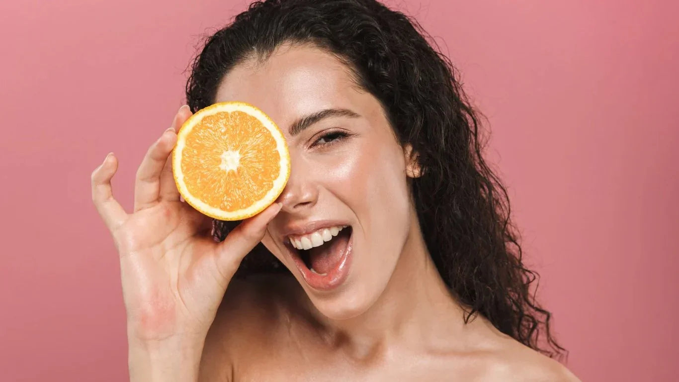 Femme souriante avec peau éclatante tenant une tranche d'orange devant un œil, fond rose